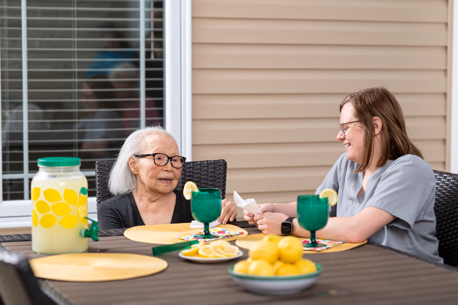 An elderly woman and a younger woman sitting at an outdoor table with a pitcher of lemonade, green glasses with lemon slices, and a bowl of lemons, enjoying a conversation on a patio with beige siding and a window in the background.