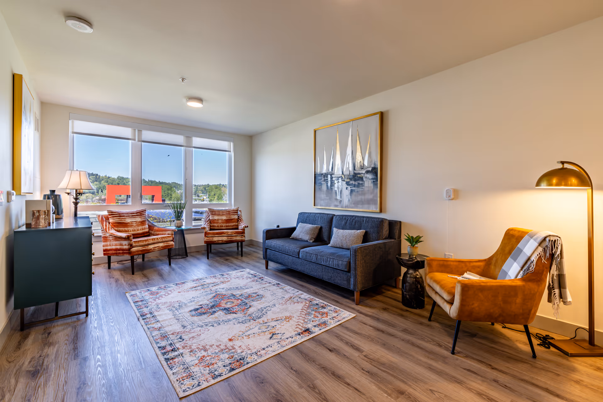 A bright modern living room with a sofa, armchairs, an area rug, large windows, and decorative artwork and lighting.