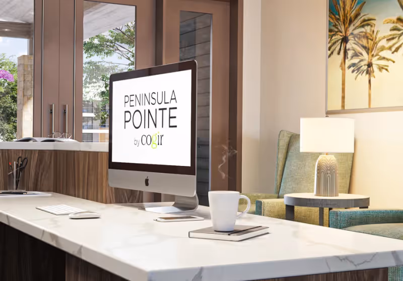 Modern reception desk with an iMac displaying "PENINSULA POINTE", a steaming coffee mug on the counter, and lounge chairs with a lamp in the background.