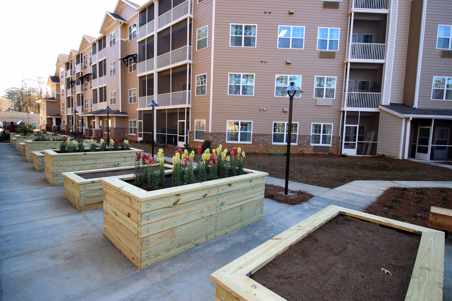 Outdoor garden area at Mulligan Park Gracious Retirement Living featuring multiple raised wooden planter boxes with some containing blooming flowers and others filled with soil. The garden is adjacent to a multi-story residential building with balconies and numerous windows. Concrete walkways and lamp posts are also visible.