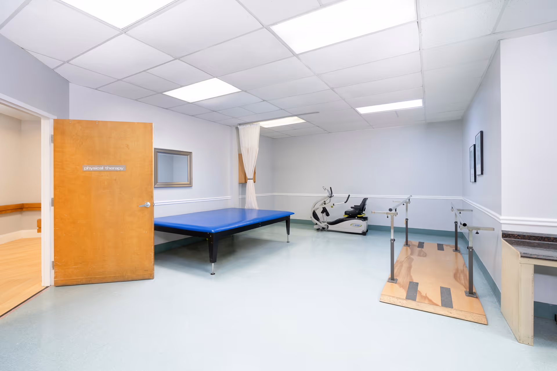 A physical therapy room with a wooden door labeled 'physical therapy' open to a hallway. Inside the room, there is a blue padded therapy table, a stationary exercise bike, and a wooden ramp with handrails for rehabilitation exercises. The room has light blue flooring, white walls with a chair rail, and a drop ceiling with fluorescent lighting.