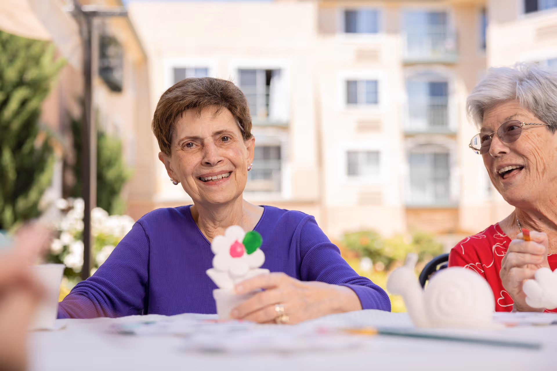 Two elderly women sitting at a table outdoors, engaging in a craft activity with painted ceramic figures, smiling and enjoying each other's company with a residential building in the background.