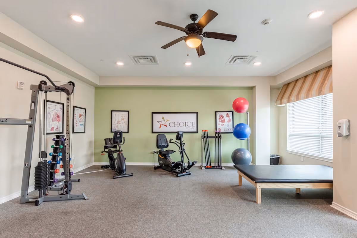 A rehabilitation exercise room with various fitness equipment including a multi-function weight machine, two stationary recumbent bikes, a rack with colorful exercise balls, and a padded exercise table. The walls are light green and beige, decorated with anatomical posters and a framed sign that reads 'Choice Rehab'. A ceiling fan with lights is mounted on the white ceiling, and a large window with blinds and a striped valance allows natural light into the room.