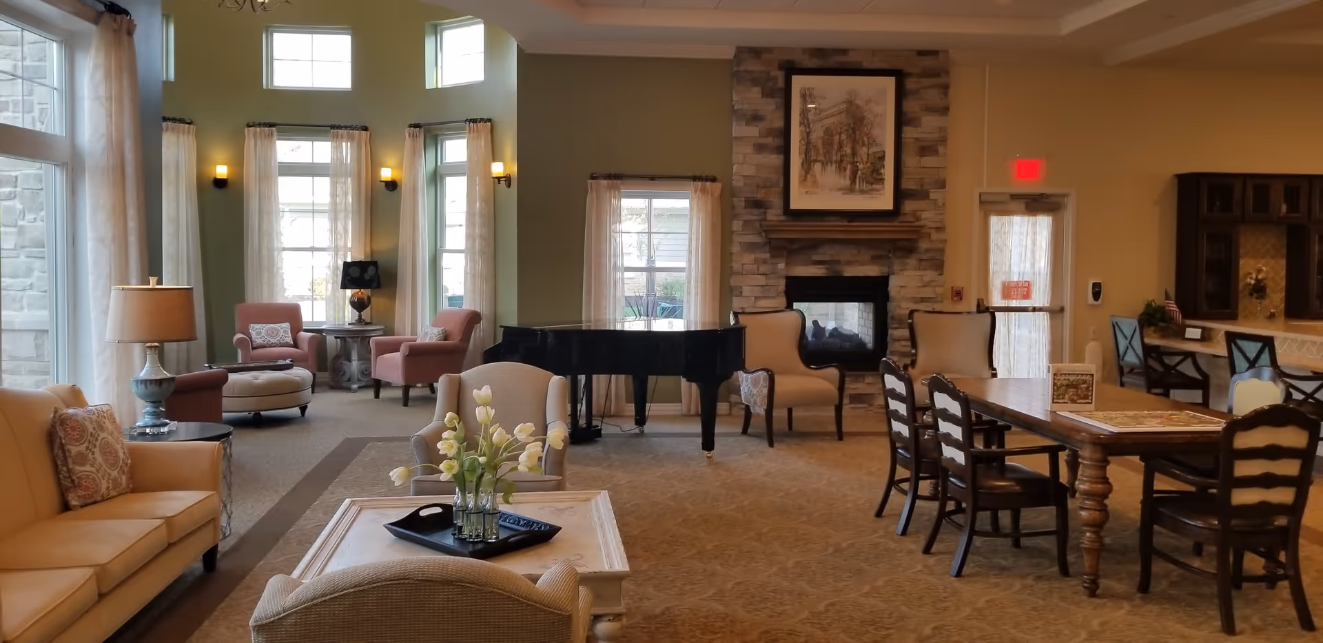 A spacious and well-lit living room area with large windows covered by sheer curtains, beige sofas and armchairs, a coffee table with a vase of white tulips, a grand piano near the windows, a stone fireplace with a framed painting above it, and a wooden dining table with chairs. The room has a warm and inviting atmosphere with soft lighting from wall sconces and lamps.