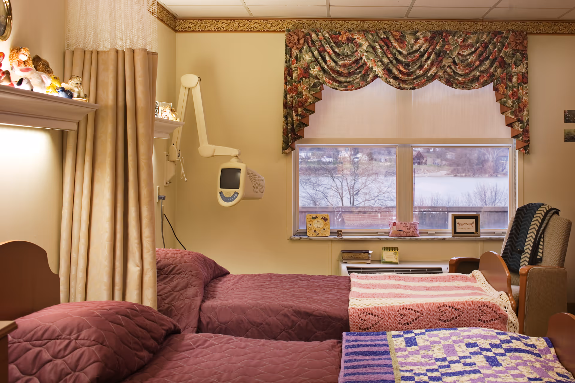 A cozy bedroom in a senior living facility with two beds covered in quilted blankets and crocheted throws. There is a window with floral valance curtains overlooking a lake and trees outside. A comfortable armchair with a blanket draped over it is positioned near the window. The room has soft yellow walls, a medical device mounted on the wall, and shelves with stuffed animals and framed pictures.