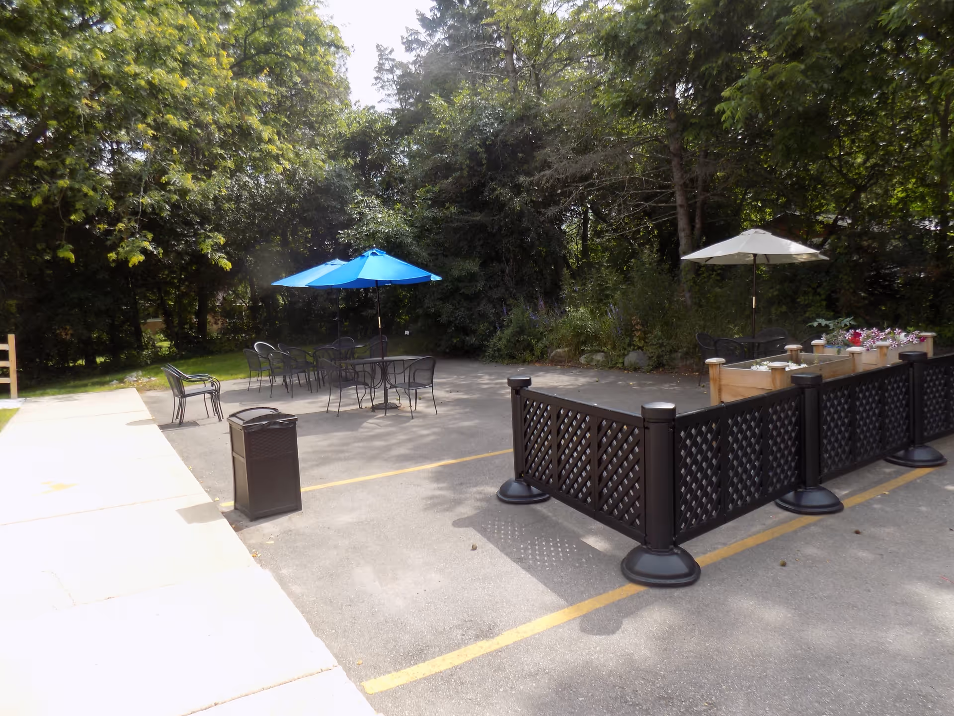 Outdoor patio area with several black metal tables and chairs, two umbrellas (one blue and one white), a trash bin, and a fenced garden bed with flowers. The area is surrounded by trees and greenery.
