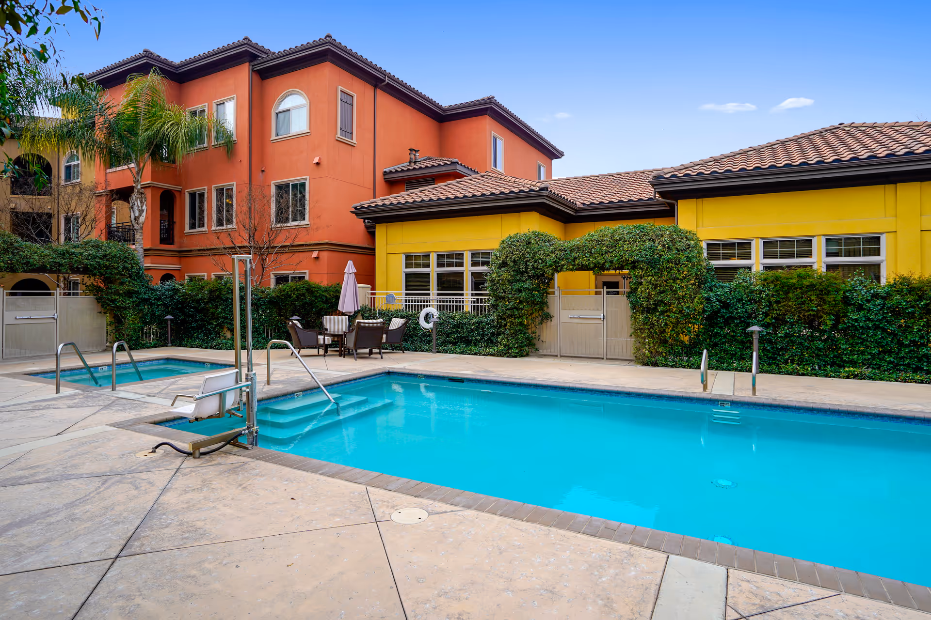 Outdoor swimming pool area at a senior living facility with a smaller hot tub adjacent to the pool. There are patio chairs and a table with an umbrella near the pool. The buildings surrounding the pool have a Mediterranean style with orange and yellow walls, tiled roofs, and greenery including palm trees and bushes.
