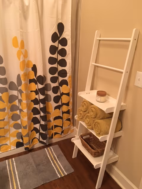 A bathroom corner featuring a white ladder-style shelf holding rolled yellow towels, a small basket, and a candle. Next to the shelf is a shower curtain with a pattern of yellow, black, and gray leaf shapes. A gray bath mat with yellow and white stripes is on the wooden floor.