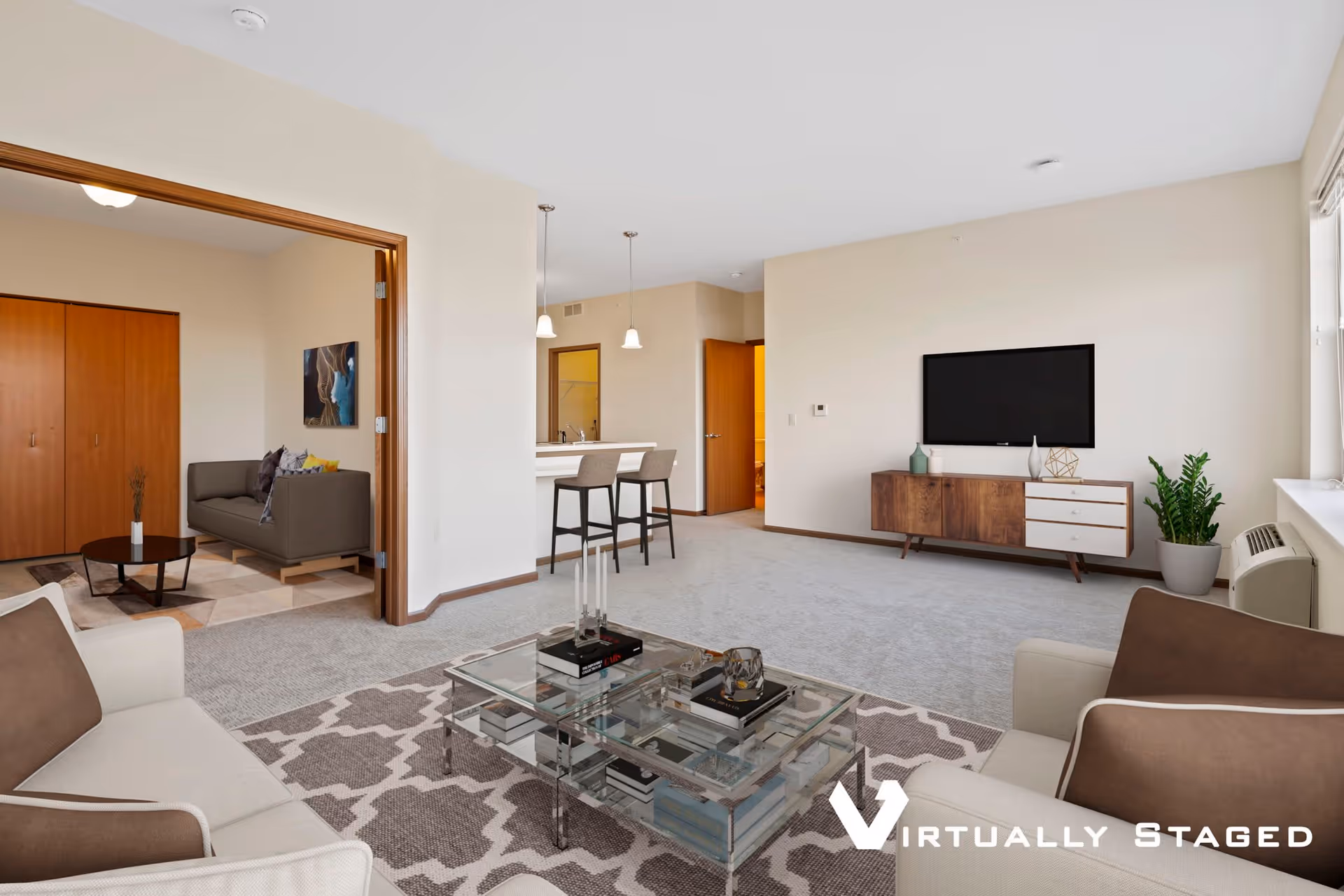 A spacious living room with beige walls and carpeted floor. The room features a glass coffee table on a patterned rug, two armchairs with brown cushions, a wooden TV stand with a flat-screen TV mounted above it, and a potted plant near the window. In the background, there is a kitchen area with two bar stools at a counter and a doorway leading to another room with a gray sofa and wooden closet doors.