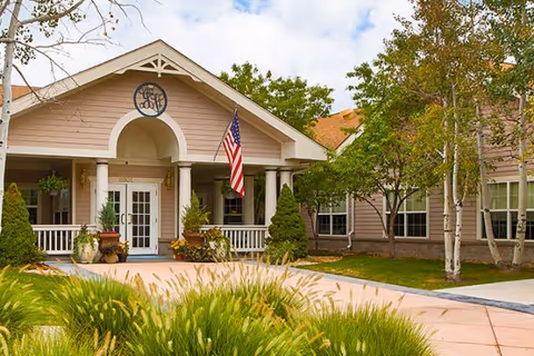 Front exterior view of Juniper Village at Aurora facility with a covered entrance, American flag, potted plants, and surrounding trees and greenery under a partly cloudy sky.