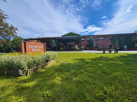 Exterior view of Thompson House Rehabilitation & Nursing Center, showing a two-story brick building with a covered entrance, surrounded by green grass and trees under a partly cloudy blue sky. A wooden sign with the facility's name is visible on the left side of the image.
