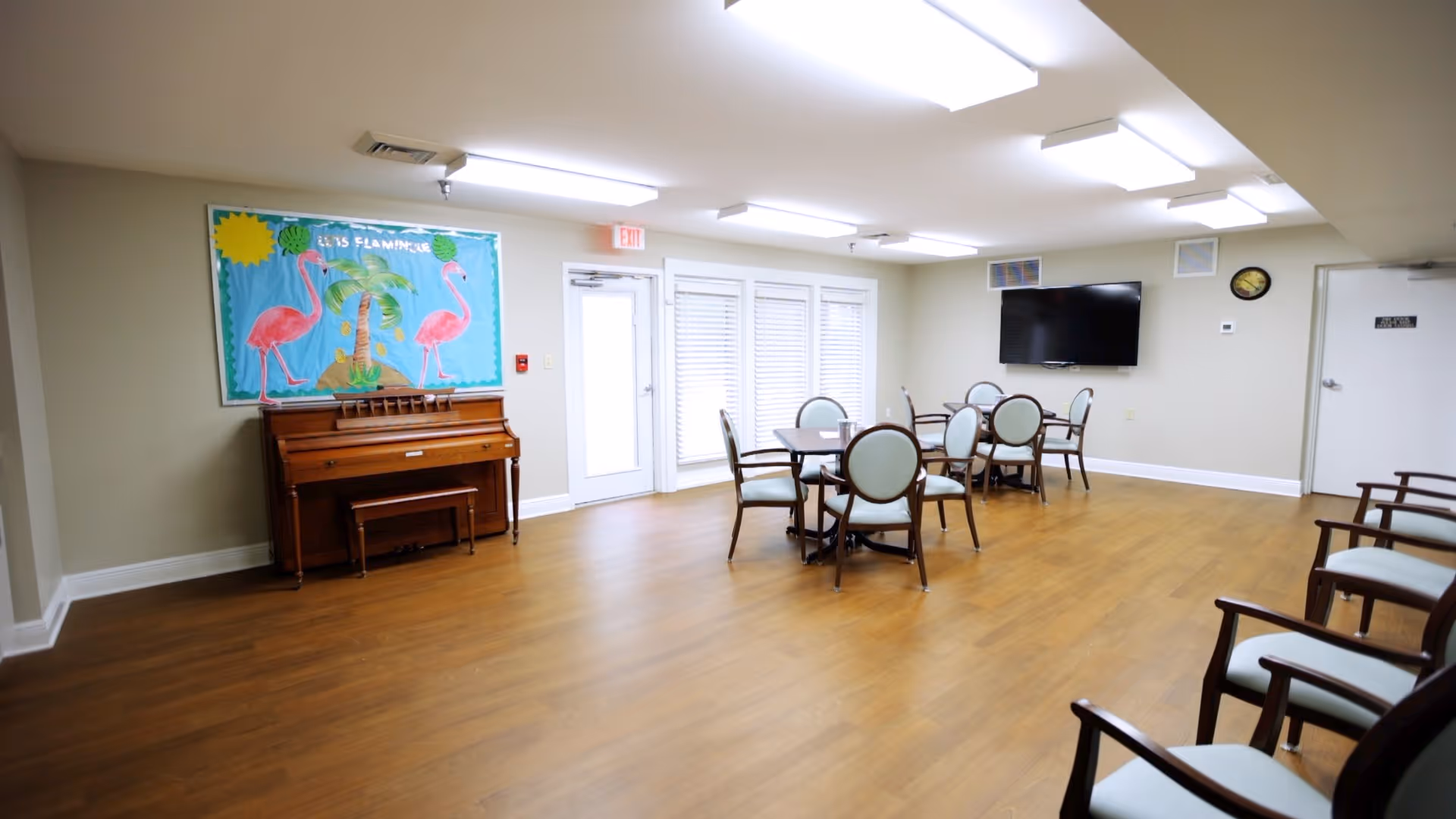 A spacious room with wooden flooring featuring several round tables surrounded by chairs with light-colored cushions. On the left wall, there is a wooden piano beneath a colorful bulletin board decorated with flamingos and a palm tree. The far wall has a set of glass doors with blinds, an exit sign above, and a flat-screen TV mounted on the right wall. Additional chairs are lined up along the right side of the room.