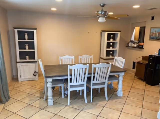 A dining room with a rectangular wooden dining table surrounded by six white wooden chairs. The room has tiled flooring and beige walls. There are two white display cabinets with decorative items on either side of the wall behind the table. A ceiling fan with a light is mounted above the table. To the right, there is a small counter opening to another room and a black refrigerator with some items on top.