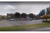Brick healthcare building with a parking lot, several cars, and trees under a cloudy sky.