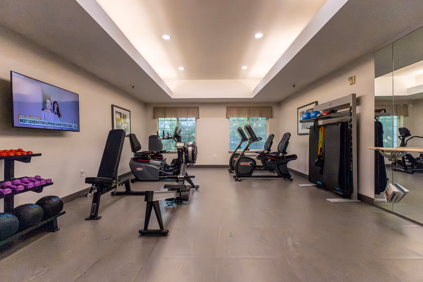 A fitness room with exercise equipment including stationary bikes, a weight bench, dumbbells, and resistance bands. The room has a large mirror on one wall, two windows with blinds, framed artwork, and a wall-mounted TV displaying a program.
