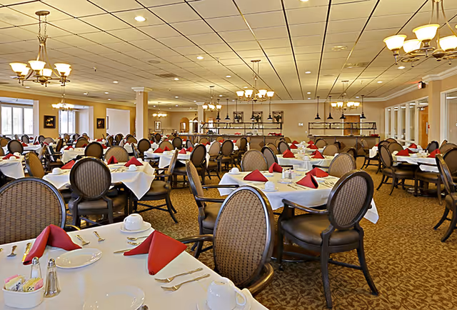A spacious dining room in a senior living facility with multiple round tables covered with white tablecloths, each set with plates, cups, silverware, and red folded napkins. The room has patterned carpet, cushioned chairs, and several chandeliers hanging from a tiled ceiling. Large windows allow natural light to enter the room.