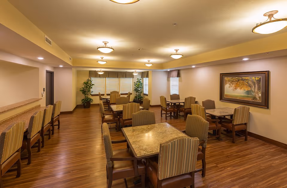 A well-lit dining room with multiple square tables and cushioned chairs arranged neatly. The room has wooden flooring, beige walls, and several ceiling lights. There are large windows with blinds and two potted plants near the windows. A framed painting of a tree is hung on one wall.