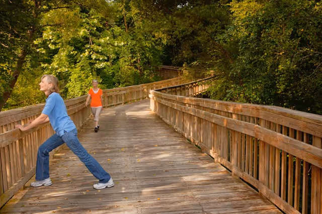 Two women on a wooden boardwalk surrounded by lush green trees. One woman in a blue shirt and jeans is stretching by leaning against the railing, while the other woman in an orange shirt and beige pants walks towards her.