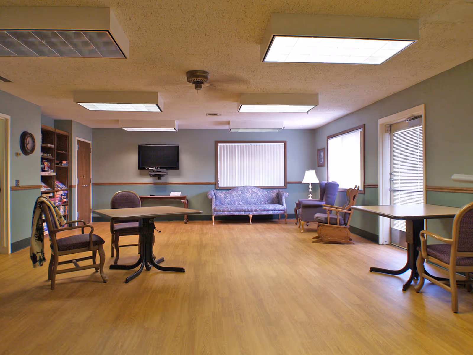 A spacious room with wooden flooring and light blue walls featuring a blue patterned couch, two armchairs, two tables with chairs, a wall-mounted TV, a lamp on a side table, and a door with blinds. The ceiling has multiple rectangular fluorescent lights.