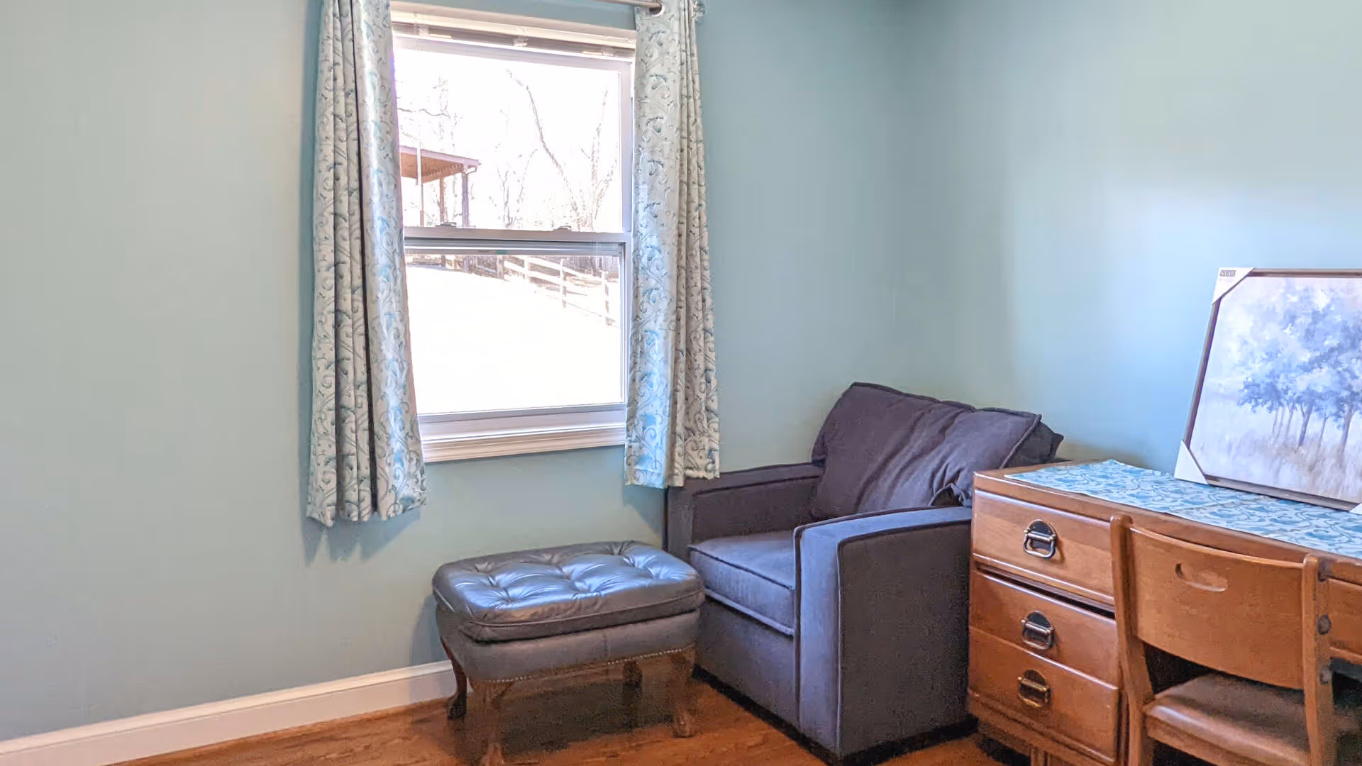 A cozy corner of a room with a blue armchair and matching ottoman placed near a window with patterned curtains. Next to the armchair is a wooden dresser with a chair and a framed painting resting on top. The walls are painted light blue.