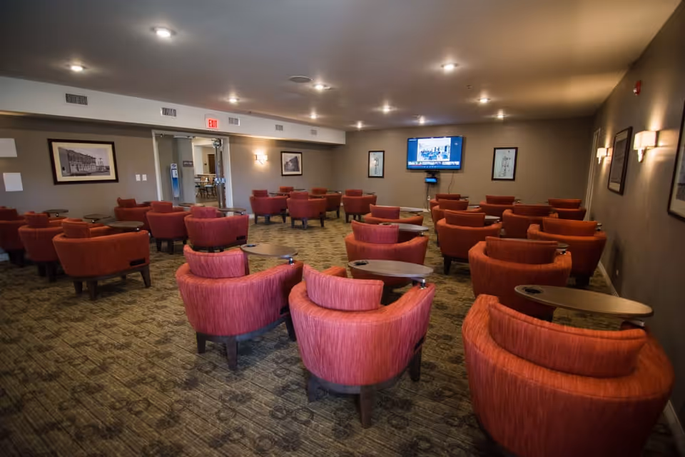 A spacious common area with multiple red upholstered armchairs arranged in rows facing a wall-mounted television. The room has carpeted flooring, neutral-colored walls with framed pictures, and ceiling lights providing illumination. There is a double glass door at the back leading to another room.