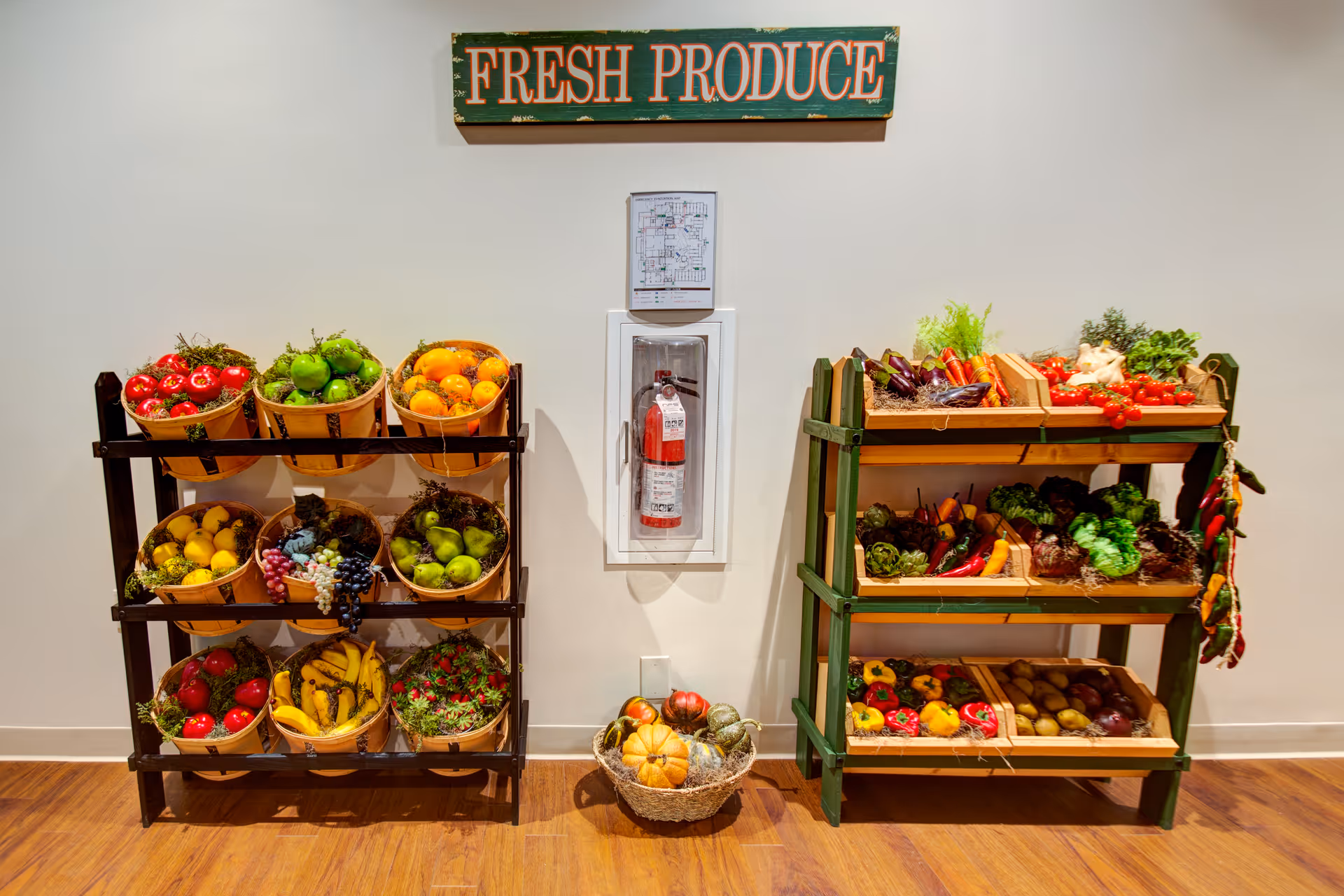 Two wooden racks filled with baskets and trays of various fresh fruits and vegetables, including tomatoes, bananas, grapes, peppers, and leafy greens, displayed against a white wall with a 'FRESH PRODUCE' sign above and a fire extinguisher mounted in the center.