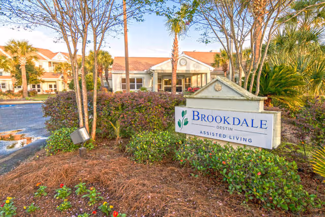 Exterior view of Brookdale Destin Assisted Living facility with a landscaped garden in the foreground, featuring bushes, flowers, palm trees, and a sign displaying the facility's name. The building has a light-colored facade with a red roof and multiple windows.
