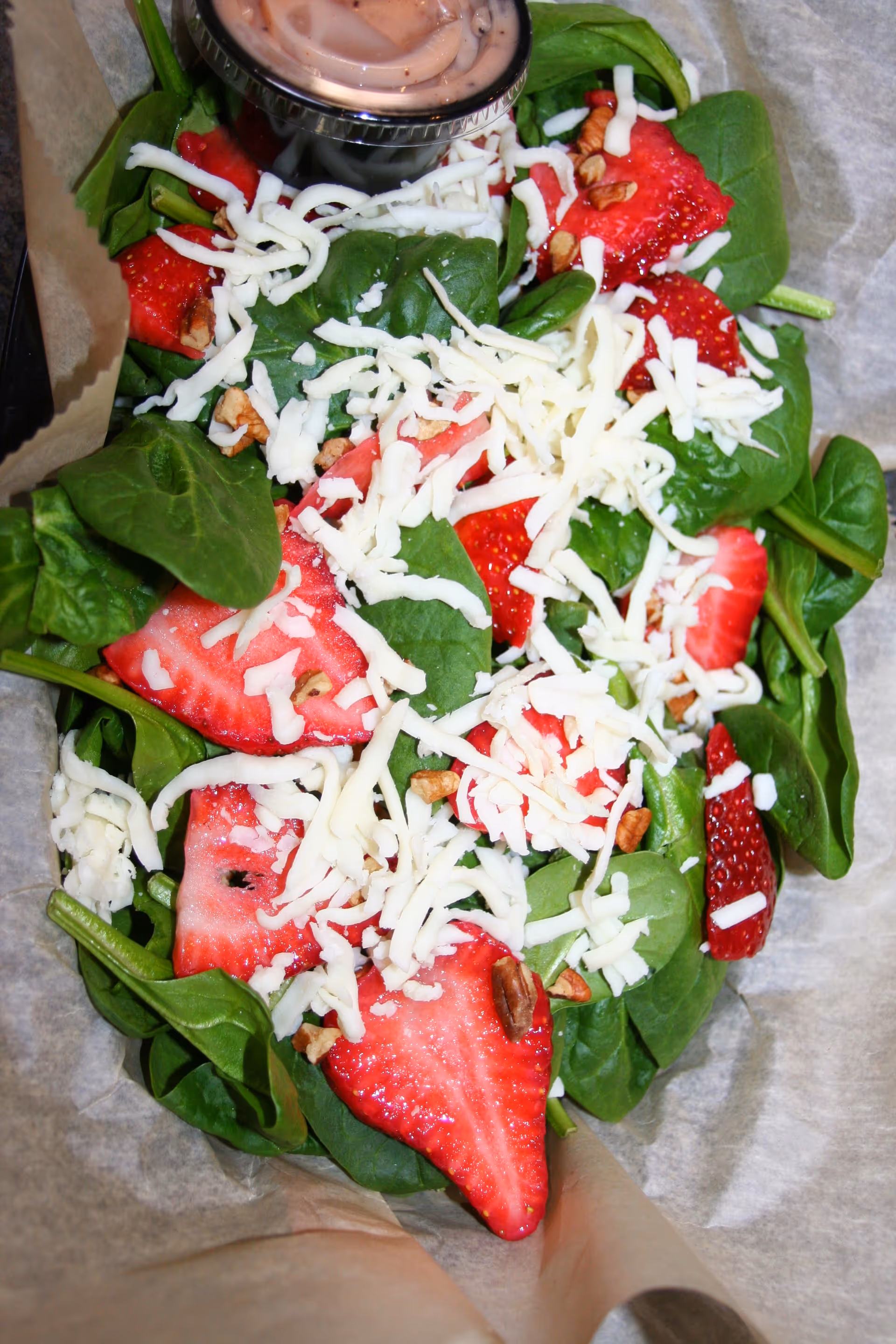 Close-up of a spinach salad topped with sliced strawberries, shredded cheese, nuts, and a small container of dressing.