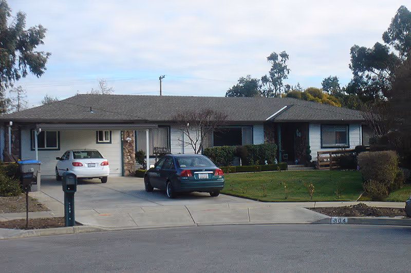Single-story residential building with a driveway containing two parked cars, a green lawn, and some bushes and trees around the property under a partly cloudy sky.