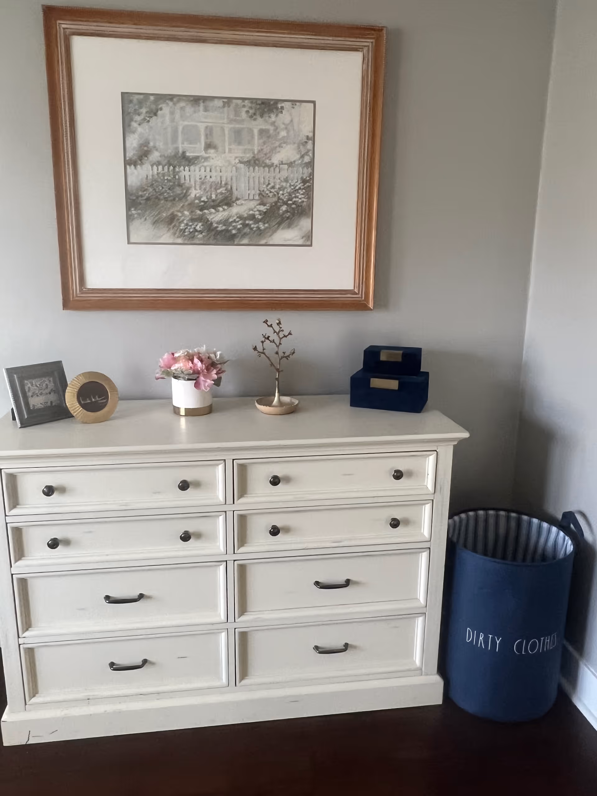 A white dresser with eight drawers against a light gray wall. On top of the dresser are two framed pictures, a small vase with pink flowers, a decorative branch in a small dish, and two stacked navy blue boxes. To the right of the dresser is a navy blue laundry basket labeled 'DIRTY CLOTHES'. Above the dresser hangs a framed painting of a garden scene with a white picket fence.