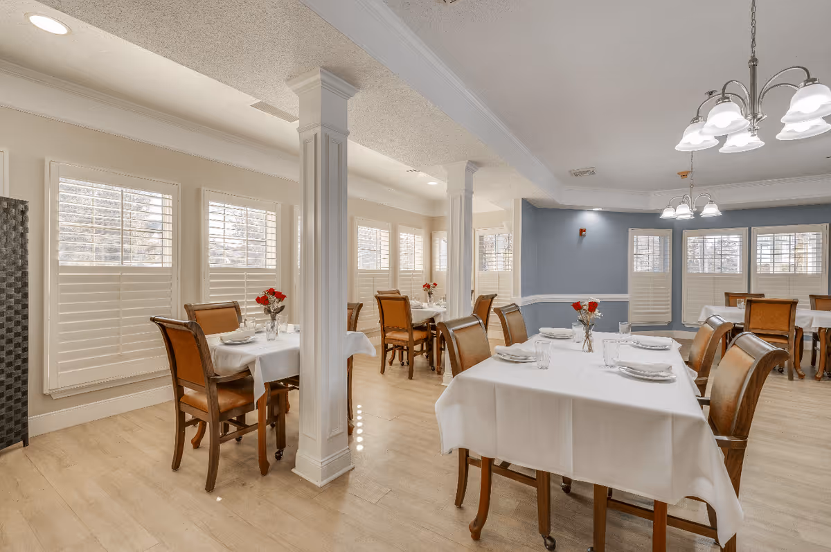 A bright dining room with several tables covered in white tablecloths, each set with plates, glasses, and napkins. The room features wooden chairs with cushioned seats, large windows with white shutters, light wood flooring, and white columns. There are small vases with red flowers on each table and ceiling light fixtures providing illumination.