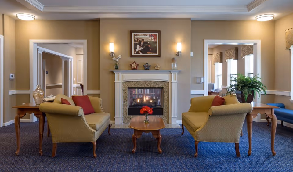 A cozy living room area with two beige upholstered armchairs facing each other across a wooden coffee table with a small vase of red flowers. Behind the chairs is a white fireplace with a lit fire, decorated with three star ornaments and a framed painting above it. The room has beige walls, blue carpet, and side tables with decorative items. There are doorways on either side of the fireplace leading to other rooms.