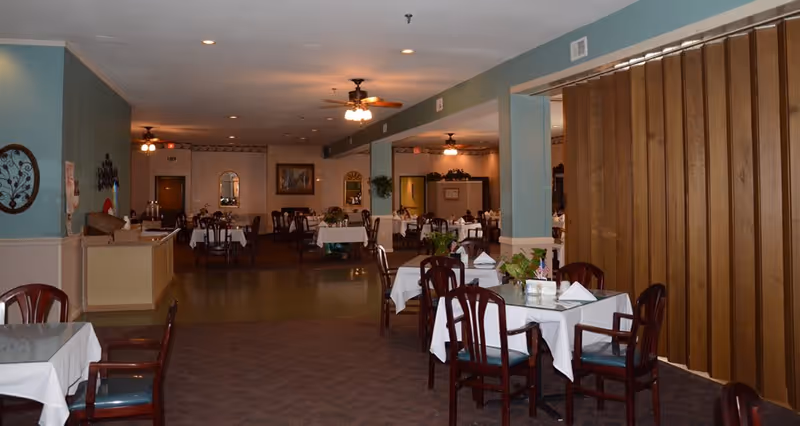 Interior view of a dining room in a senior living facility with multiple tables covered with white tablecloths and wooden chairs. The room has ceiling fans with lights, decorative wall mirrors, and plants on some tables. The walls are painted in light blue and beige tones, and there is a wooden partition on the right side.