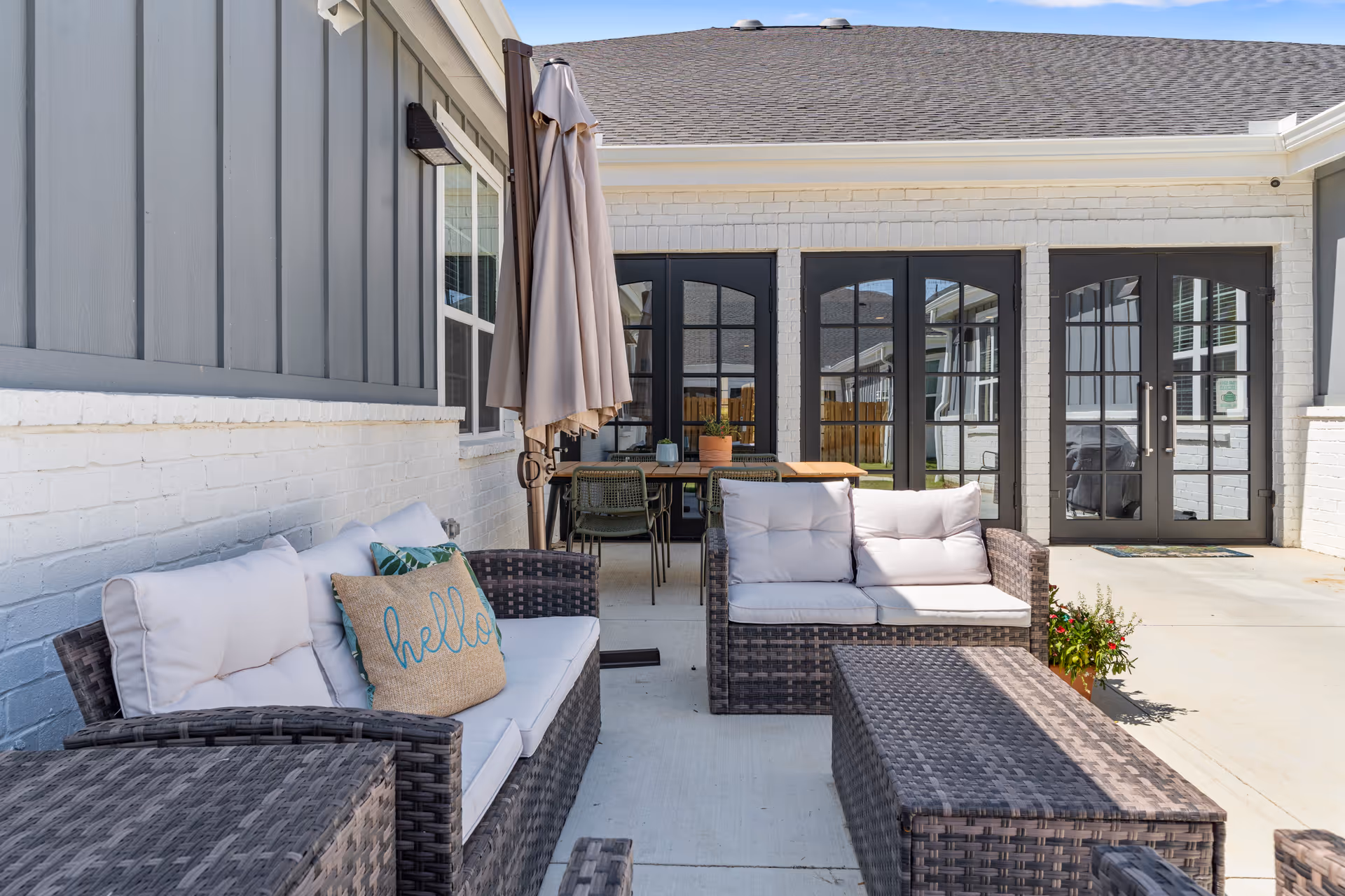 Outdoor patio area with wicker furniture including cushioned sofas and a coffee table, a closed beige umbrella, a wooden table with chairs, and glass doors leading inside a building.