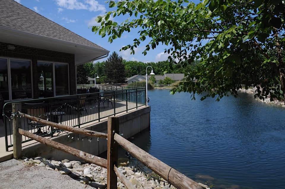 Outdoor view of a patio area next to a calm pond with a wooden fence and trees. The patio has tables and chairs and is attached to a building with large windows. The sky is clear with a few clouds.