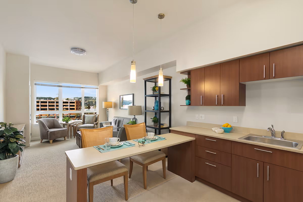 Interior view of a senior living facility apartment at Merrill Gardens at Auburn featuring a kitchen area with wooden cabinets, a countertop with a sink, and a small dining bar with two chairs set with dishes and glasses. In the background, there is a living area with a sofa, two armchairs, a floor lamp, a shelving unit with decorative items, and large windows showing an outdoor view.