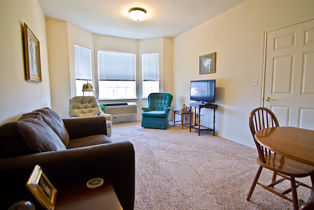 Sunlit living room with a sofa, two armchairs, a TV on a small stand, and a round dining table near a bay window.