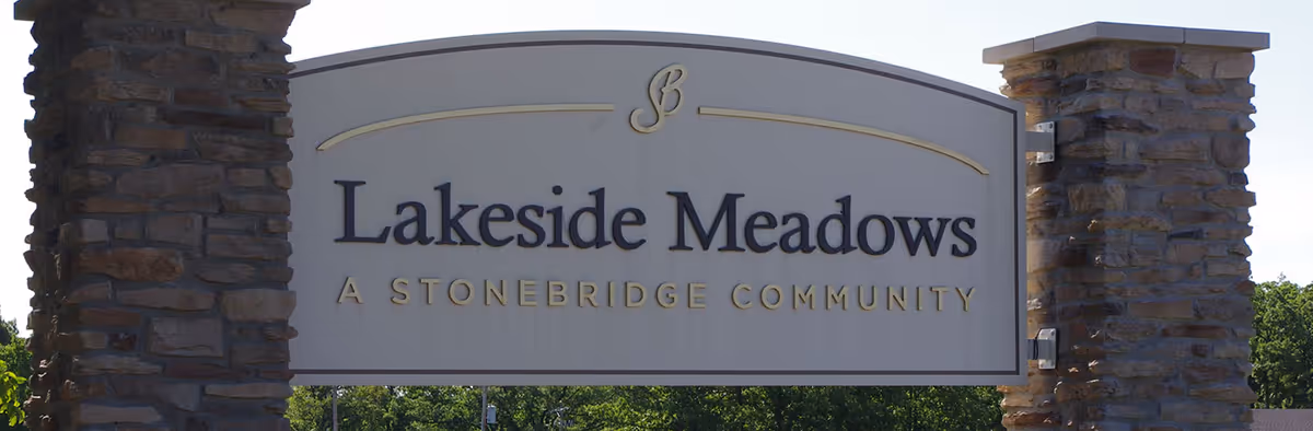 Stone pillars supporting a large sign that reads 'Lakeside Meadows A Stonebridge Community' with trees and a clear sky in the background.