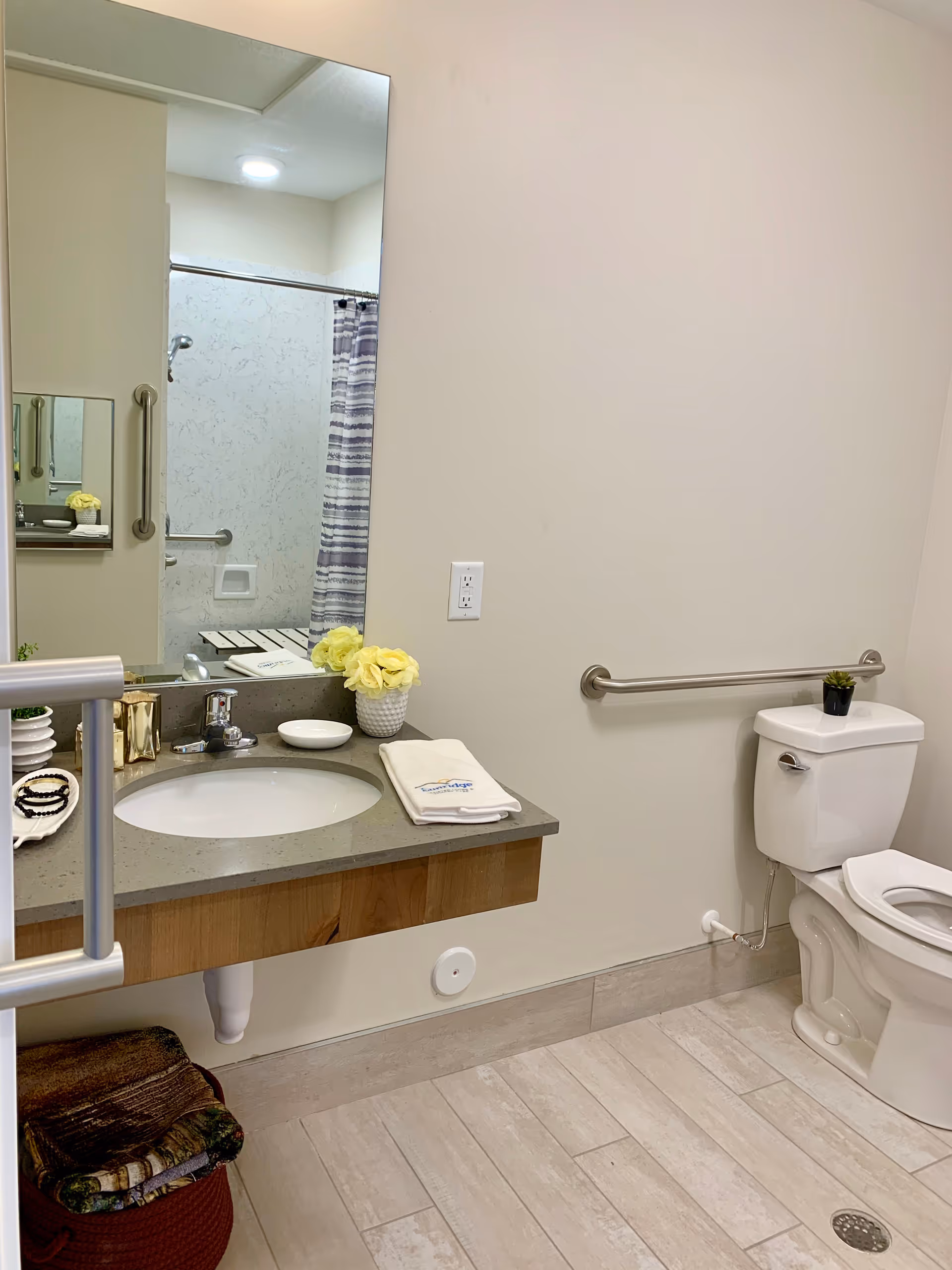 A clean and accessible bathroom with a wall-mounted sink, a large mirror, a toilet with a grab bar on the wall, and a shower area with a striped shower curtain visible in the mirror. The bathroom has light-colored walls and tiled flooring, with a small potted plant on the toilet tank and a vase with yellow flowers on the sink counter.