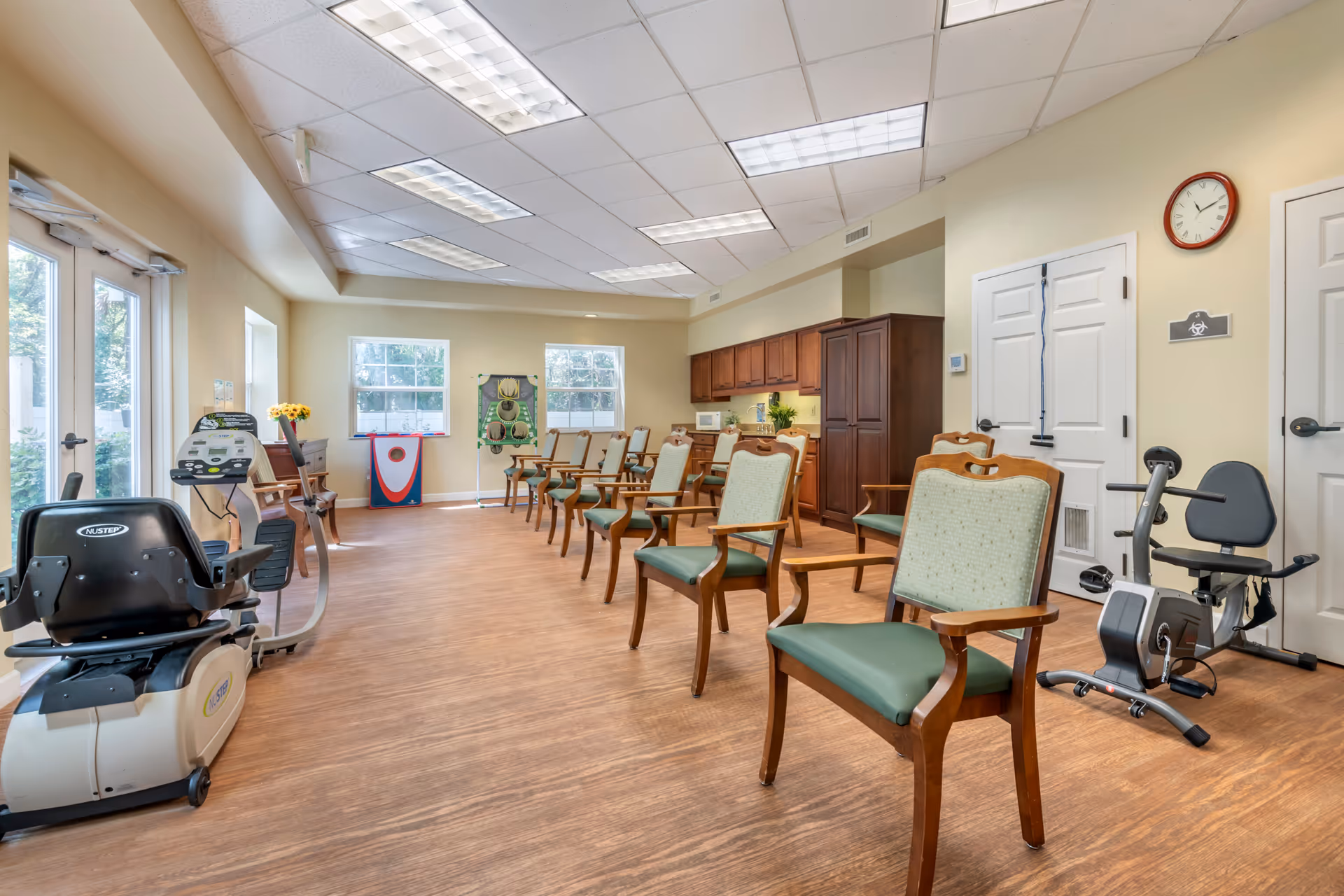 A spacious room with wooden flooring and beige walls, featuring a row of wooden chairs with green cushions arranged in a line. There are exercise machines including a recumbent bike and another stationary bike. The room has large windows letting in natural light, a clock on the wall, and a kitchenette area with wooden cabinets and a microwave. There are also some game boards and a door leading outside.