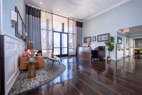 Bright modern reception lobby with tall glass entry doors, a seating area, a reception desk, decorative plants, and dark wood floors.