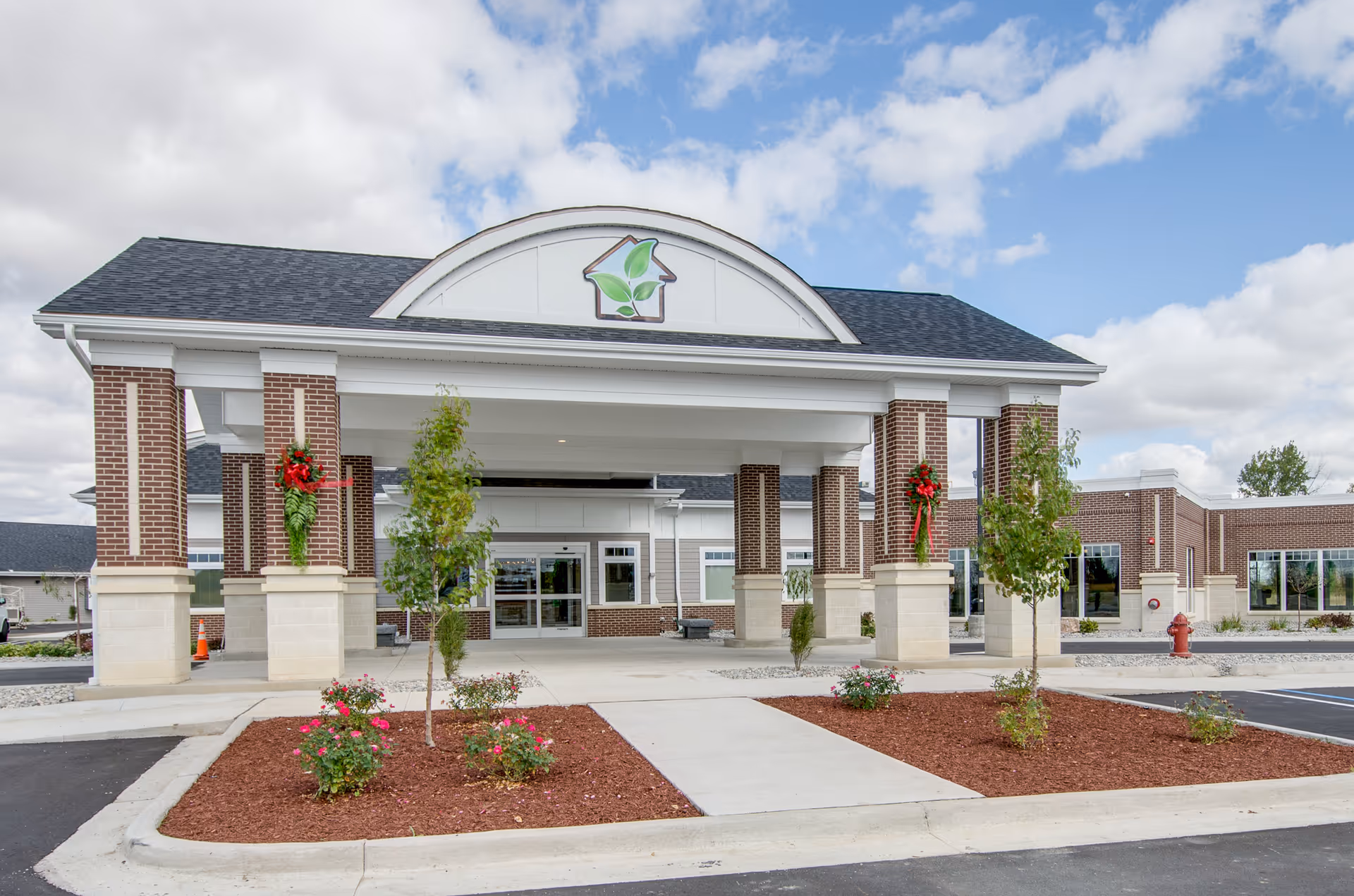 Front exterior view of Great Lakes Rehab Center showing a covered entrance with brick pillars decorated with red bows and greenery, small landscaped areas with young trees and flowers, and a clear sky with scattered clouds.