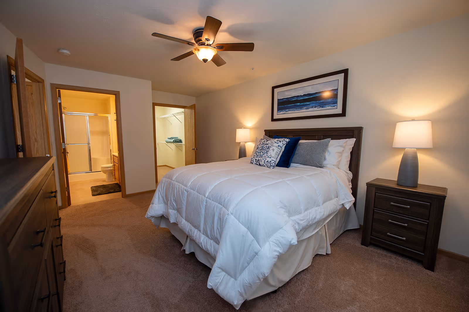 A cozy bedroom with a neatly made bed featuring white bedding and decorative pillows. The room has beige carpet, a wooden dresser on the left, and a wooden nightstand with a lamp on the right. Above the bed hangs a framed picture of a seascape. The ceiling has a fan with a light. Two open doorways reveal a bathroom with a shower and toilet on the left and a walk-in closet with shelves and towels on the right.
