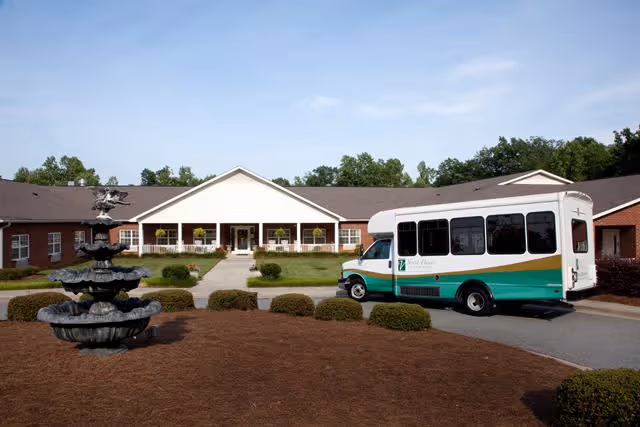 Front entrance of a single-story senior living building with a tiered fountain in the foreground and a green-and-white shuttle bus parked at the driveway.