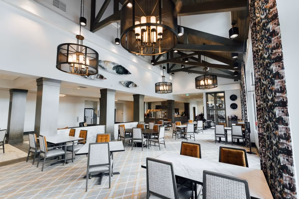Spacious dining room with multiple tables and chairs, high exposed-beam ceiling, and large pendant chandeliers.