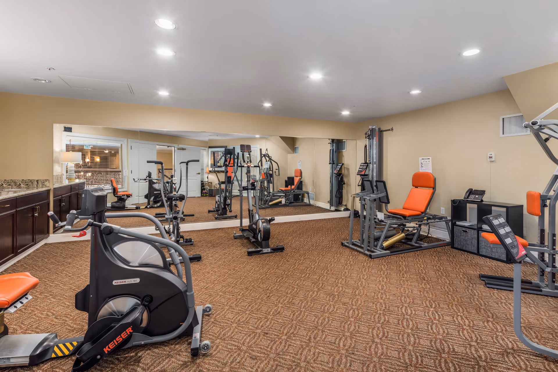 A fitness room with various exercise equipment including a stationary bike, weight machines, and a rowing machine. The room has beige walls, a large mirror on one wall, recessed ceiling lights, and brown patterned carpet. There are orange padded seats on the equipment and a countertop with cabinets along one side.