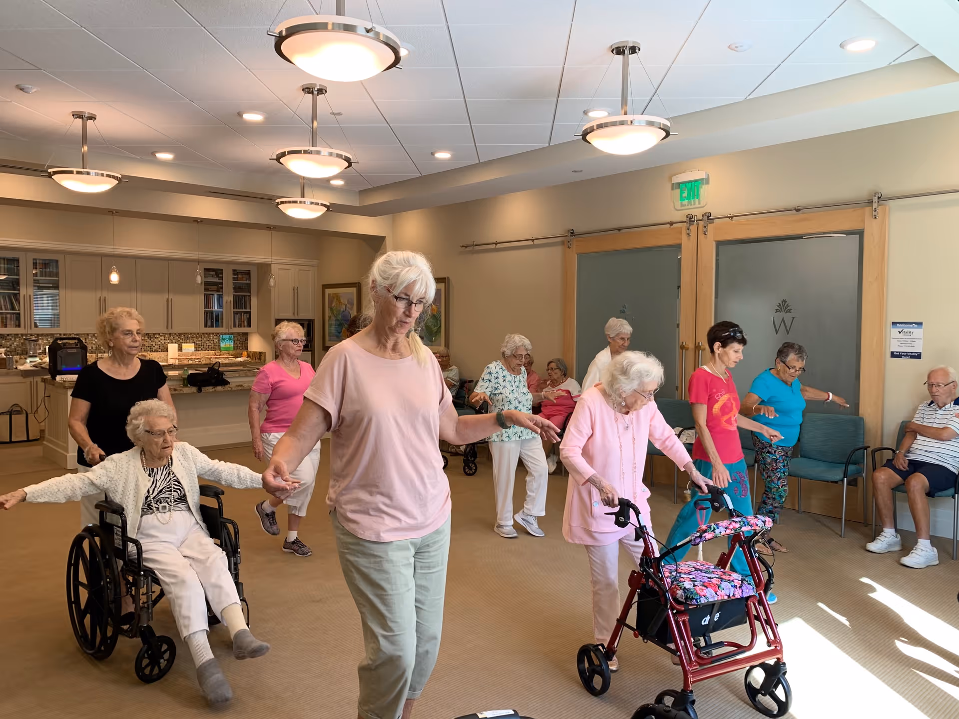 A group of elderly individuals participating in a seated and standing exercise class in a well-lit room with beige walls and carpet. Some participants use mobility aids such as a wheelchair and a walker. The room has modern ceiling lights, a kitchenette area with cabinets and a countertop in the background, and frosted glass doors with a 'W' logo.
