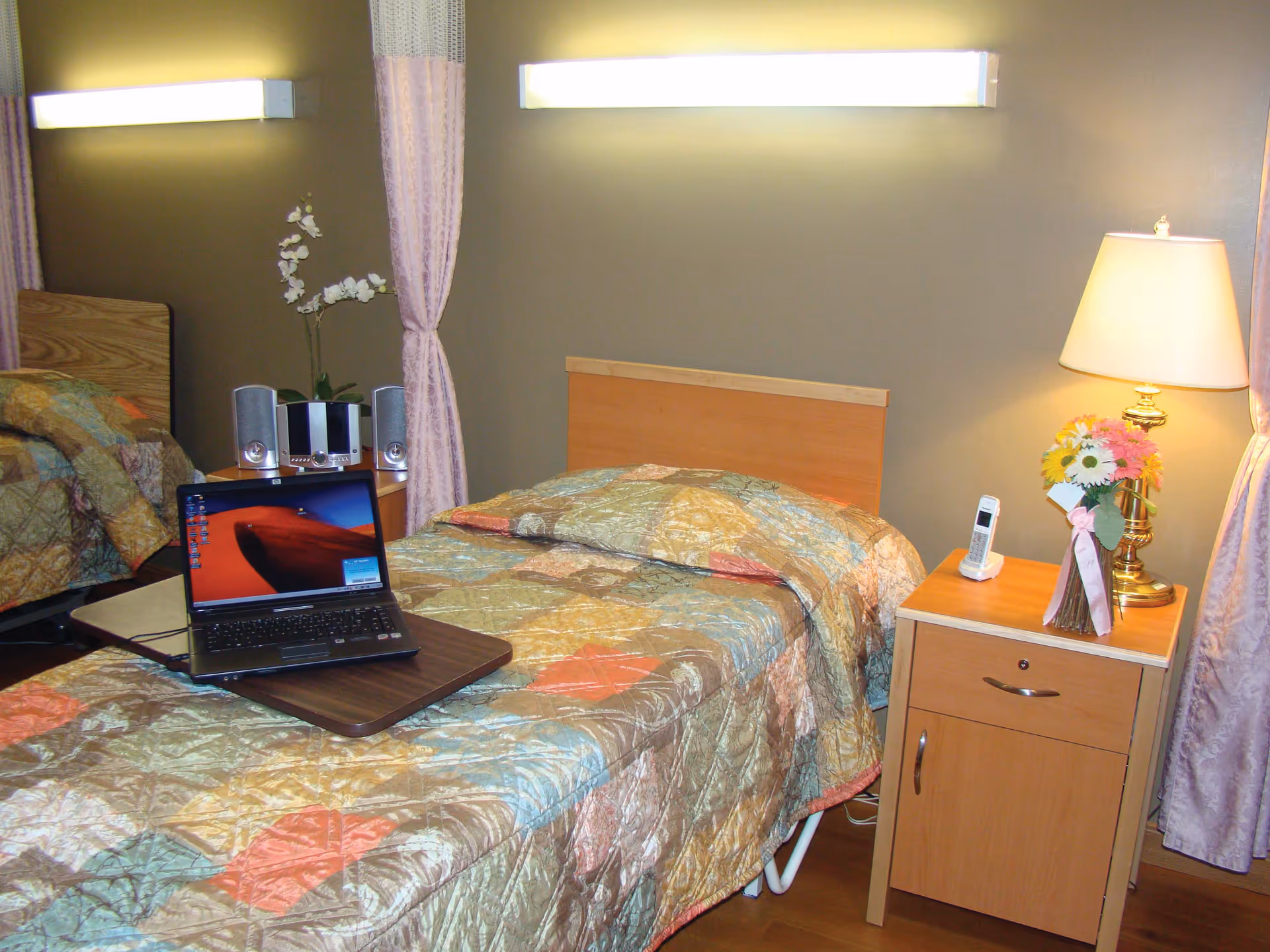 A senior living facility bedroom with a single bed covered in a colorful quilt. On the bed is a laptop on a small tray table. Next to the bed is a wooden nightstand with a lamp, a cordless phone, and a vase of flowers. The room has beige walls, two wall-mounted lights, and curtains partially drawn around the bed area.