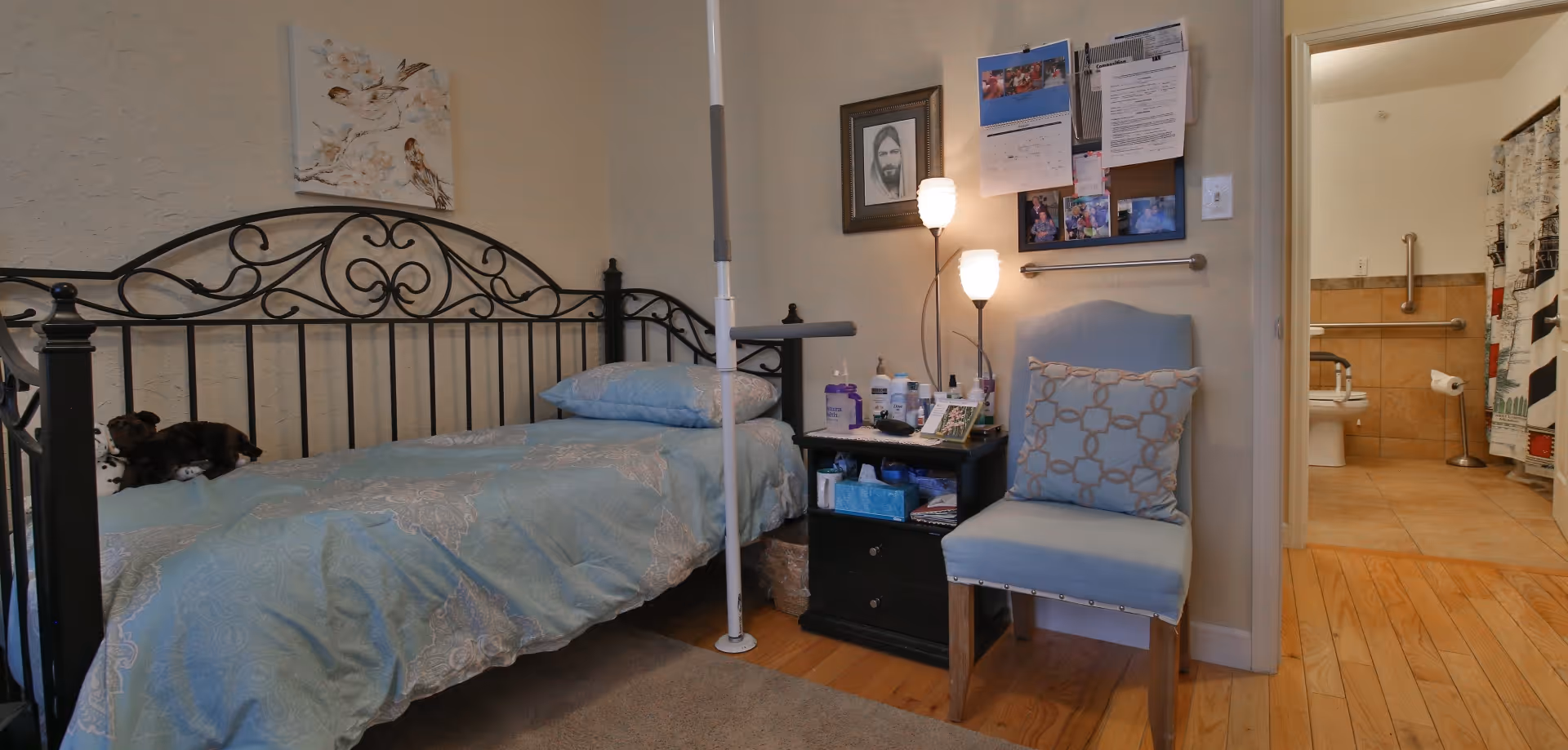 A cozy bedroom with a black metal daybed covered in a light blue patterned comforter and pillows. A small black nightstand beside the bed holds various personal items and two lit lamps. A light blue cushioned chair with a patterned pillow is next to the nightstand. On the wall above the chair are framed pictures and a bulletin board with photos and papers. The room has wooden floors and a doorway leading to a bathroom with tiled floors and grab bars near the toilet.