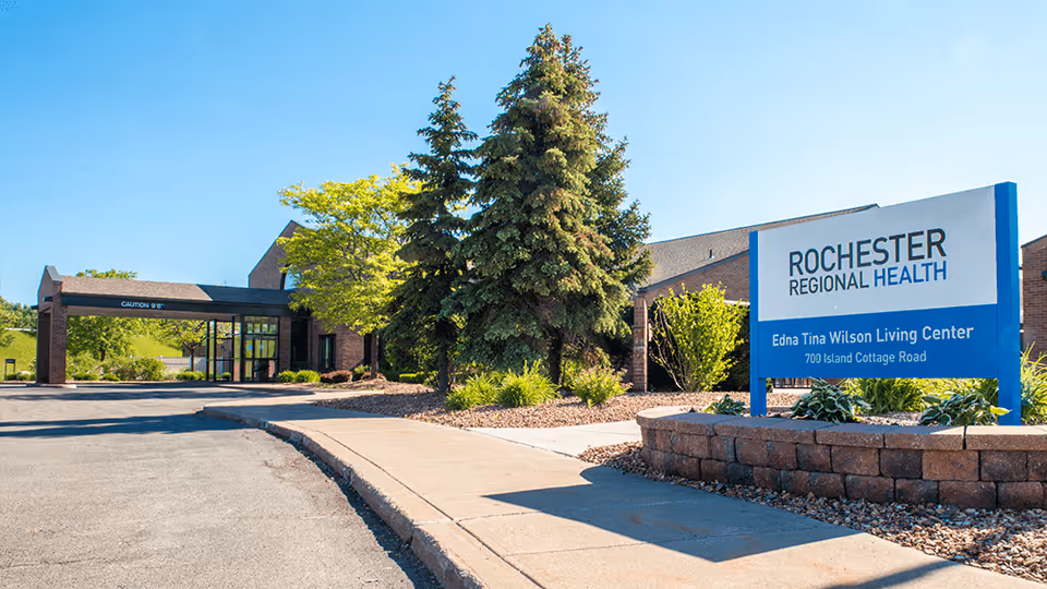 Exterior view of the Rochester Regional Health Edna Tina Wilson Living Center building with a clear blue sky, trees, and a paved driveway leading to the entrance.
